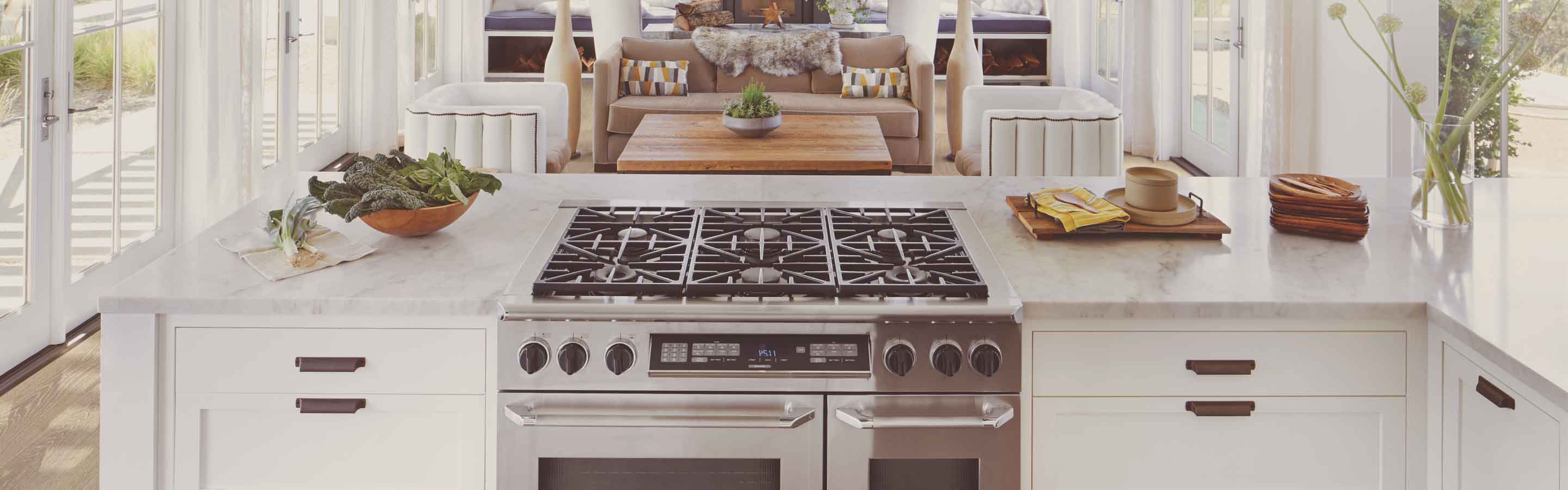 Luxury white marble kitchen island with built-in stainless steel range, styled with vegetables, wooden serveware, and flowers, set in front of a bright modern living room.
